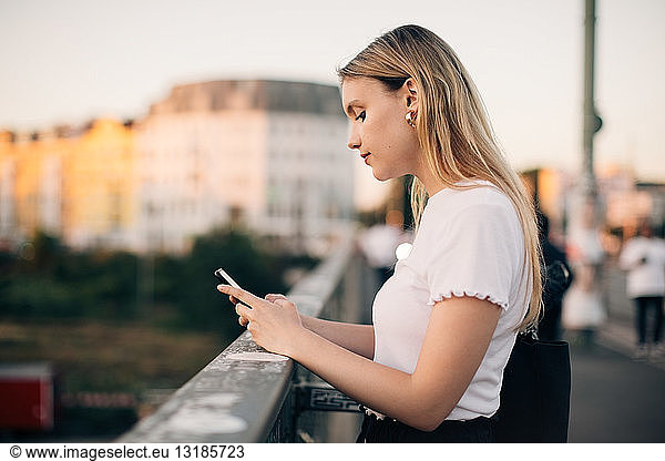 Side view of young woman using smart phone while standing on bridge in city
