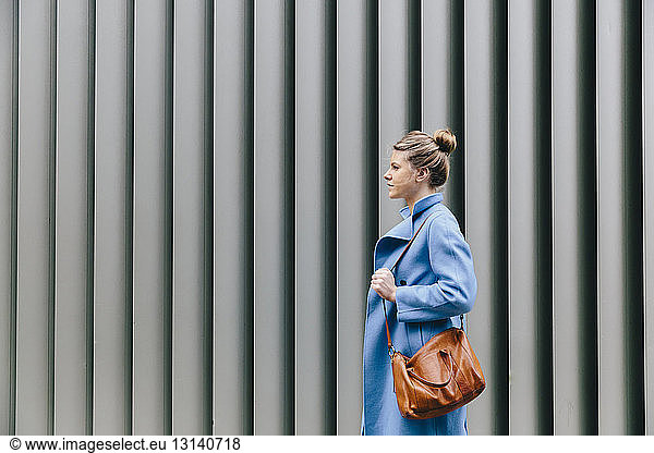Side view of young woman wearing trench coat while carrying purse against shutter in city