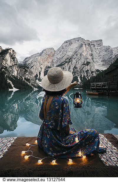 Side view of young woman with illuminated string lights and lantern sitting by lake against mountains