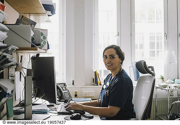 Side view portrait of female nurse sitting at desk with computer in clinic