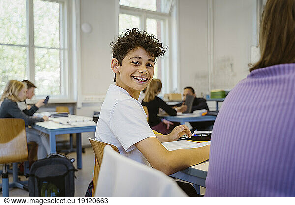Side view portrait of smiling schoolboy sitting at desk in classroom
