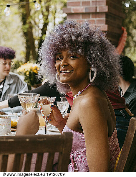 Side view portrait of smiling transwoman with curly hair sitting with friends during party in back yard