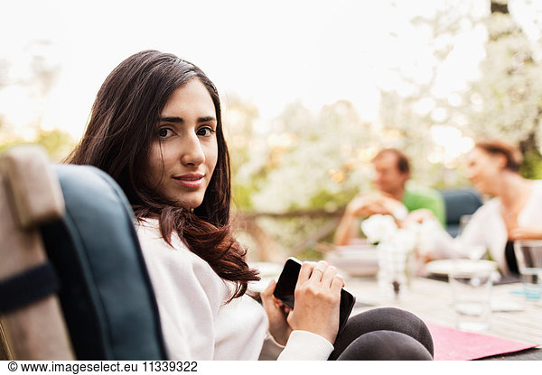 Side view portrait of teenage girl holding smart phone while parents sitting in background at yard