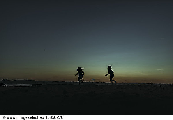 Sihouette of brother and sister running towards ocean at dusk