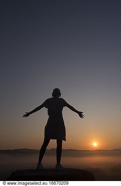 Silhouette of girl at mountain top with outstretched arms at sunrise