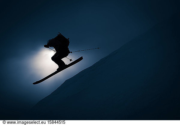 Silhouette of man going off jump with skis and backpack
