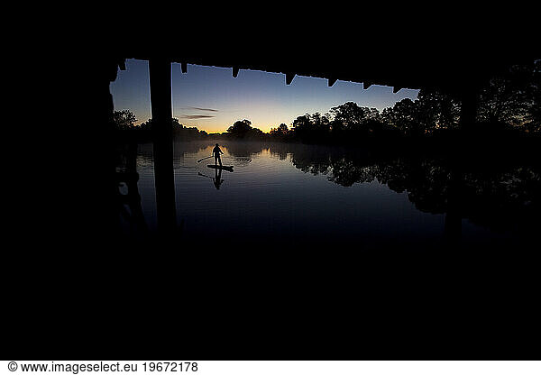 Silhouette of one man stand up paddleboarding in sunrise light with trees reflecting in the water.