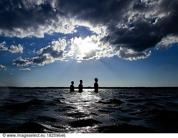 Silhouette of teenage boys in lake with sun rays and clouds