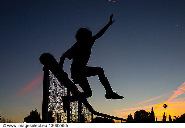 Silhouette sporty girl jumping over fence during dusk against sky