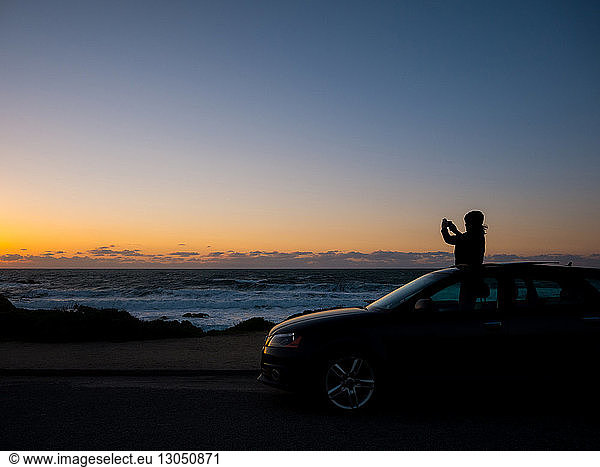 Silhouette teenage girl photographing while standing in sun roof during sunset