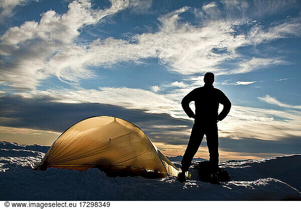 Silhouetted hiker standing next to backlit tent on mountain summit.