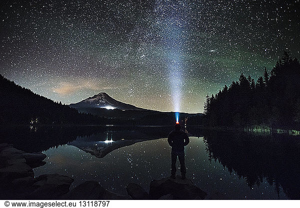 Silhouetten-Mann mit Scheinwerfer steht am Seeufer vor Sternenhimmel