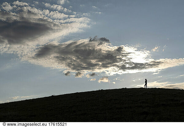 Silhouettierte Frau steht auf einem Grashügel im späten Tageslicht und beobachtet den Sonnenuntergang über der Prärie im ländlichen Saskatchewan; Saskatchewan  Kanada
