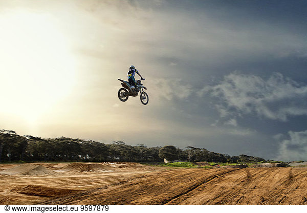 Silhouettierter junger  männlicher Motocross-Rennfahrer  der in der Luft über die Schlammbahn springt.