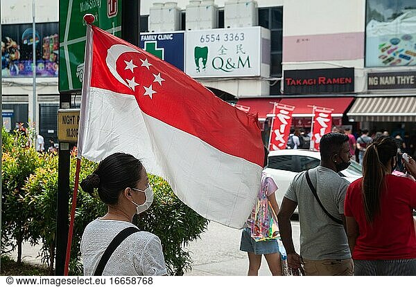 Singapur  Republik Singapur  Asien - Eine Frau trägt eine Schutzmaske  um sich und andere vor einer Infektion mit dem Coronavirus (Covid-19) zu schützen. Neben ihr ist anlässlich des Nationalfeiertags am 9. August eine Nationalflagge aufgestellt worden.