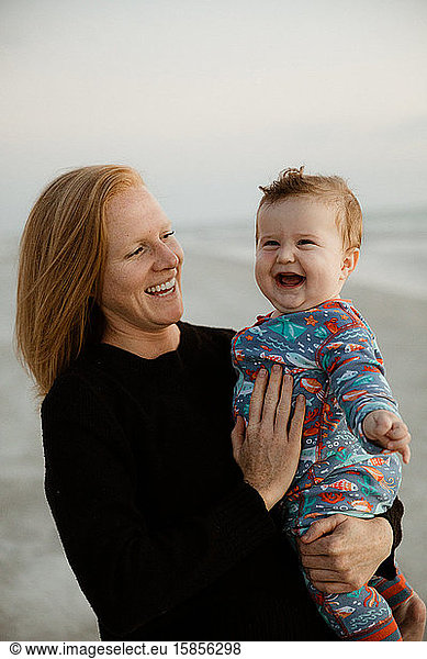 single mom with red hair holds laughing chubby baby boy on the beach