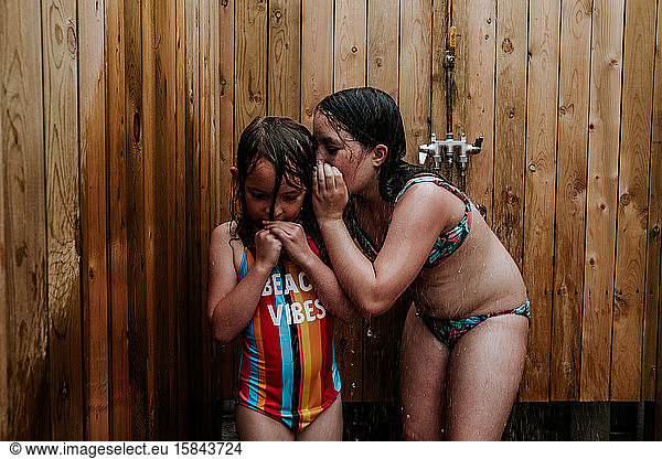 sister whispering in younger sister's ear in outdoor shower