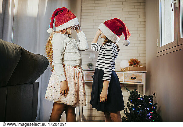 Sisters peeking at each other from Santa hats in living room