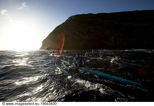 Six men in one man outrigger canoes paddling next to cliffs into the sunset.