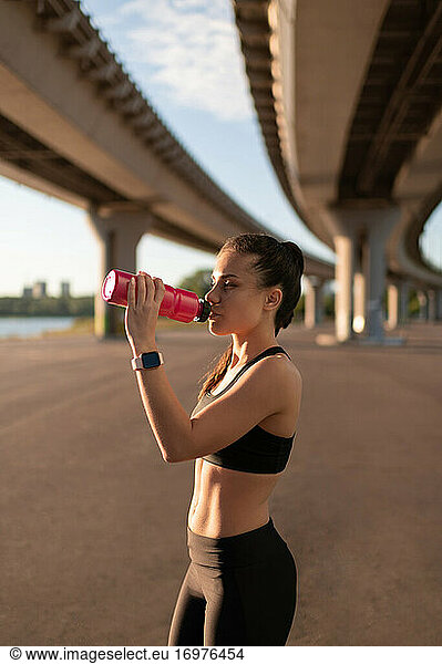 Slim sportswoman drinking water on shore under bridge