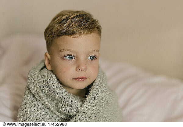 Small blonde boy with blue eyes wrapped in grey blanket sitting on bed