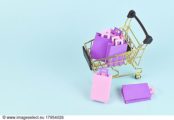 Small golden shopping cart filled with pink and purple paper shopping bags on side of blue background with copy space