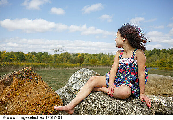 Small windblown girl with bare feet sits on boulder outside in summer