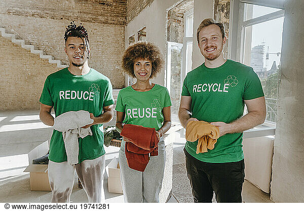 Smiling activists holding secondhand garments and standing in apartment