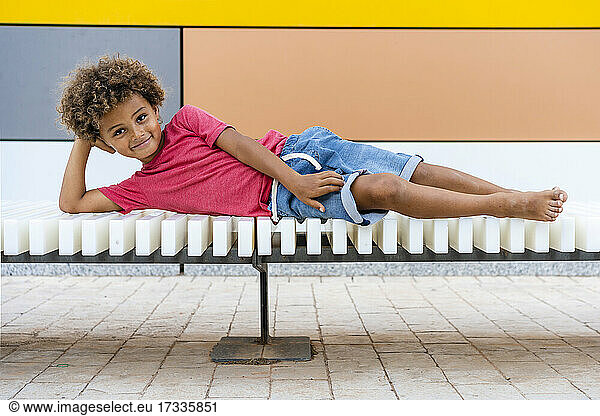 Smiling Afro boy lying on bench in front of wall