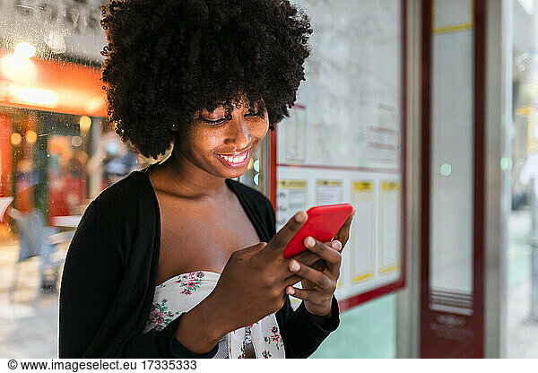 Smiling Afro woman texting message through mobile phone at bus stop