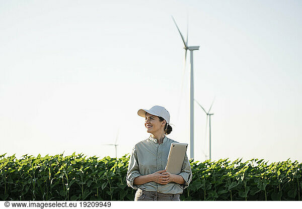 Smiling agronomist holding laptop at field with wind turbines in background