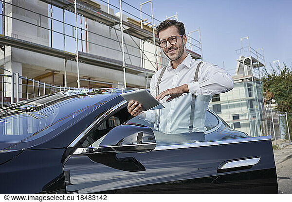 Smiling architect with tablet PC standing by car at construction site