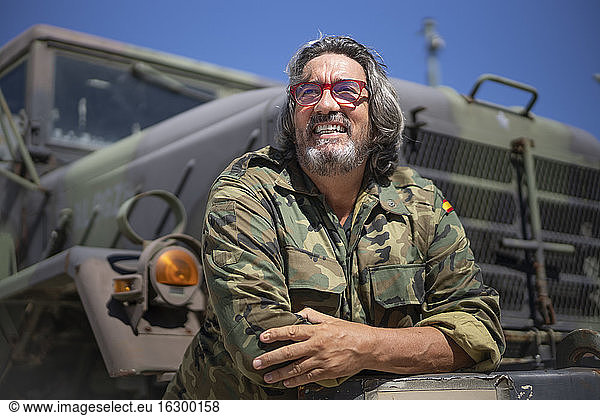 Smiling army soldier with arms crossed leaning on military truck during sunny day