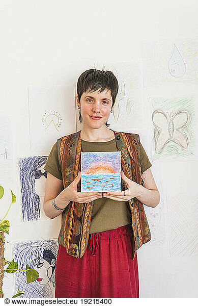 Smiling artist holding painting at wall