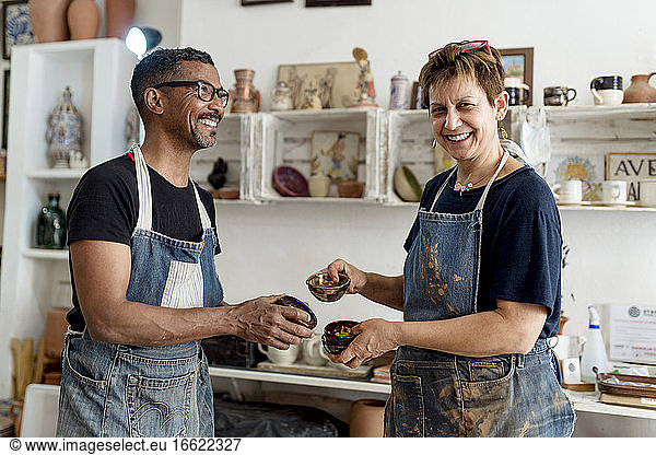 Smiling artists holding ceramics while standing in workshop