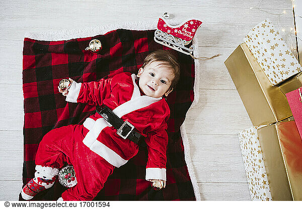 Smiling baby boy in Santa Claus costume lying on blanket at home during Christmas