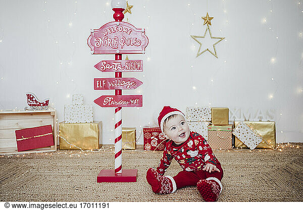 Smiling baby girl wearing Santa hat playing while sitting by direction pole at home during Christmas