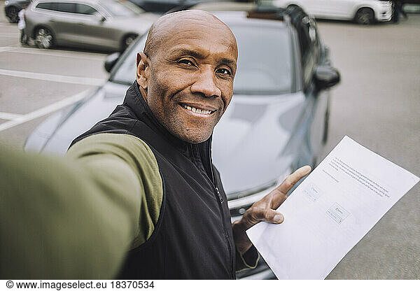 Smiling bald man showing car contract while taking selfie
