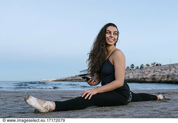 Smiling ballet dancer looking over shoulder while doing leg split at beach against clear sky