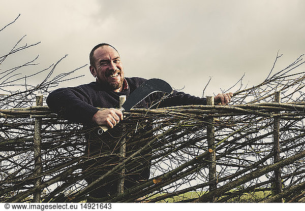Smiling bearded man holding bill hook standing next to a newly built traditional hedge.