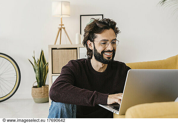 Smiling bearded man using laptop sitting by sofa at home