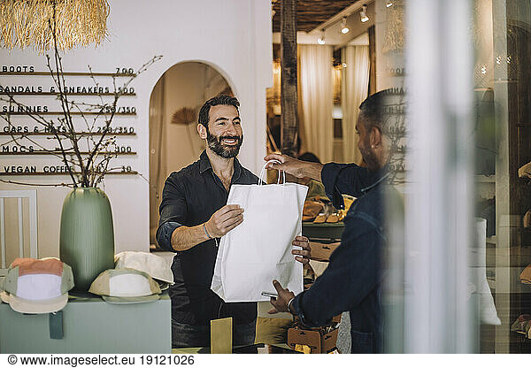 Smiling bearded salesman giving bag to male customer at boutique
