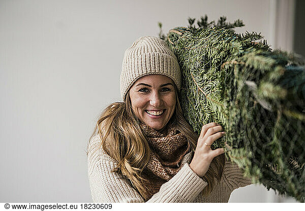 Smiling beautiful blond woman carrying Christmas tree at home
