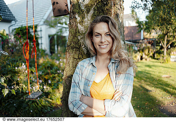 Smiling beautiful blond woman standing with arms crossed in front of tree trunk at backyard