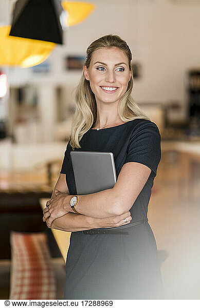 Smiling beautiful female entrepreneur holding digital tablet in office while looking away