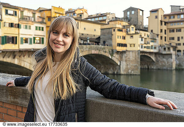 Smiling beautiful woman with bangs standing by wall in front of Ponte Vecchio Bridge