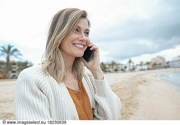 Smiling beautiful young blond woman talking on smart phone at beach
