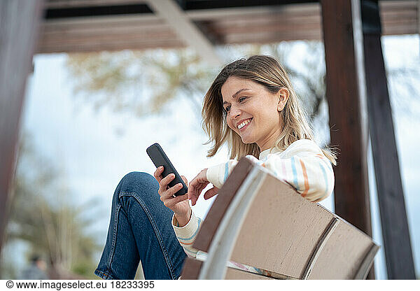 Smiling beautiful young woman sitting on bench using mobile phone