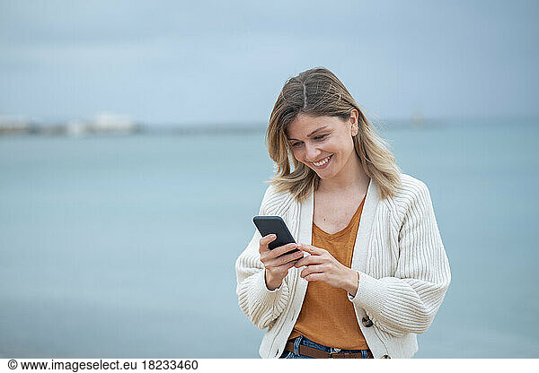 Smiling beautiful young woman text messaging on mobile phone at beach