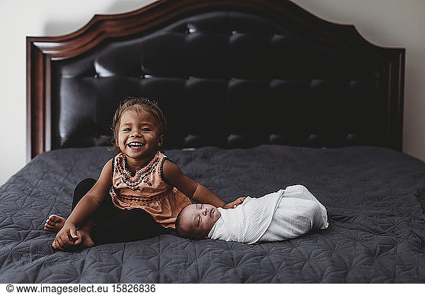 Smiling biracial 2 yr old girl on bed with infant sibling